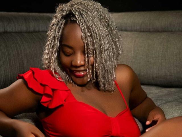 KaiaJhons with dark skin, curly grayish-white hair, and red ruffled top, smiling with eyes closed, sitting on gray couch, showing her tits in tight top.