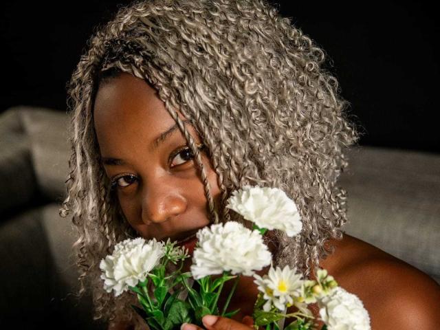 KaiaJhons with curly greyish-blonde hair, brown eyes, and dark skin, holding white chrysanthemums, partially covering her small, dark nipples. Black background.