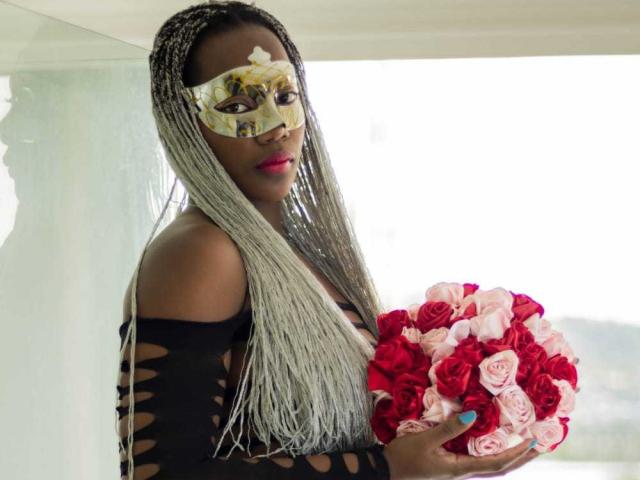 KaiaJhons with long silver braids, wearing a gold mask and black cut-out dress, holding a red and pink rose bouquet, has pink lipstick and blue nails.