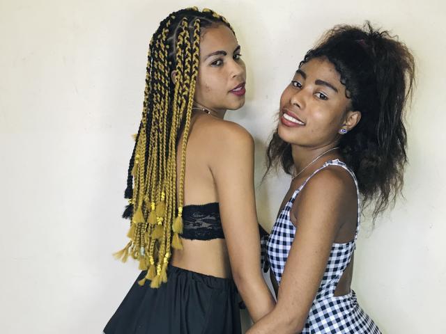 SylEtChar with yellow braids and black lace top, back to camera, holding hands with SylEtChar in checkered dress, both smiling, SylEtChar with curly hair, white wall.