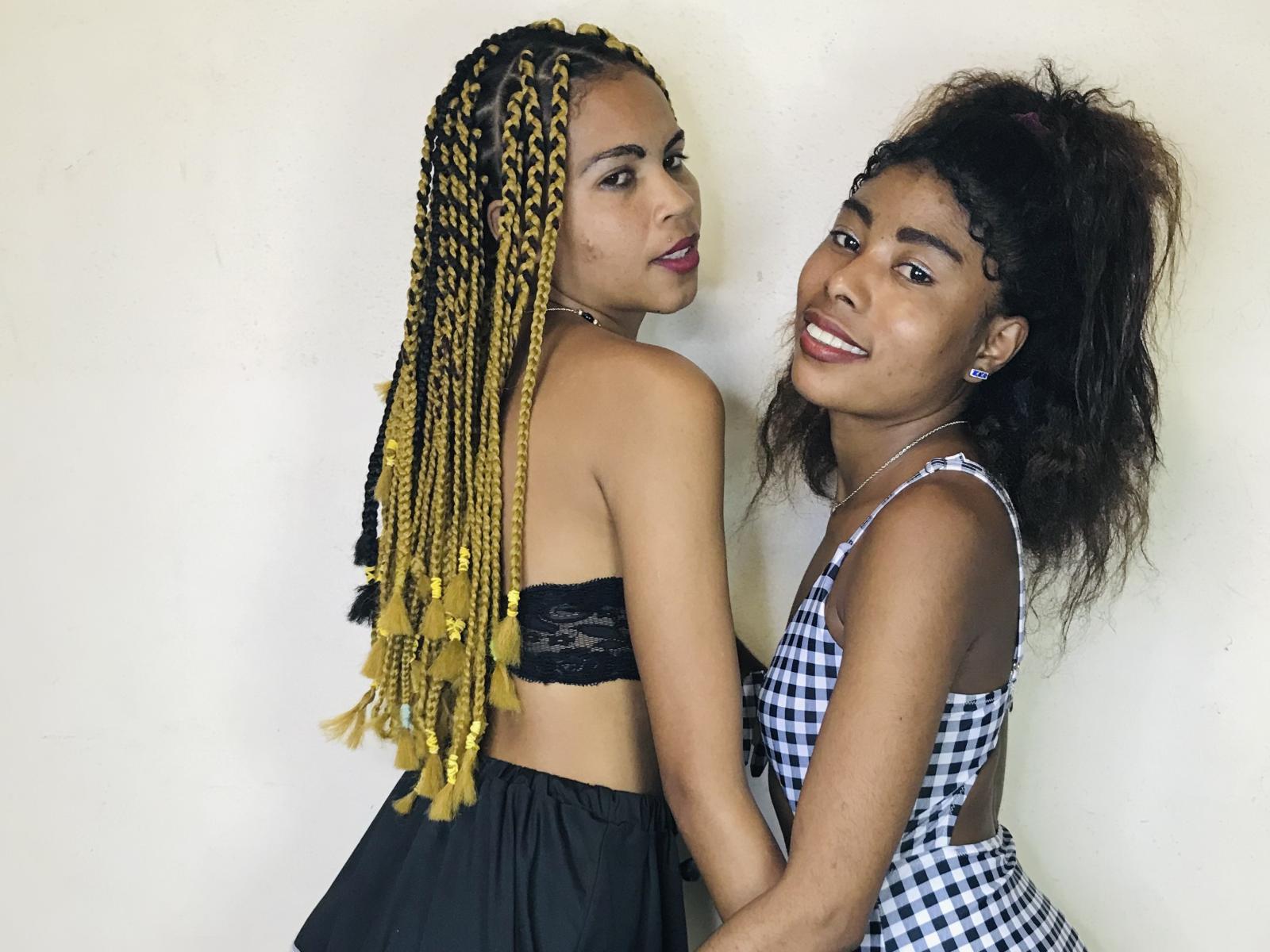 SylEtChar with yellow braids and black lace top, back to camera, holding hands with SylEtChar in checkered dress, both smiling, SylEtChar with curly hair, white wall.