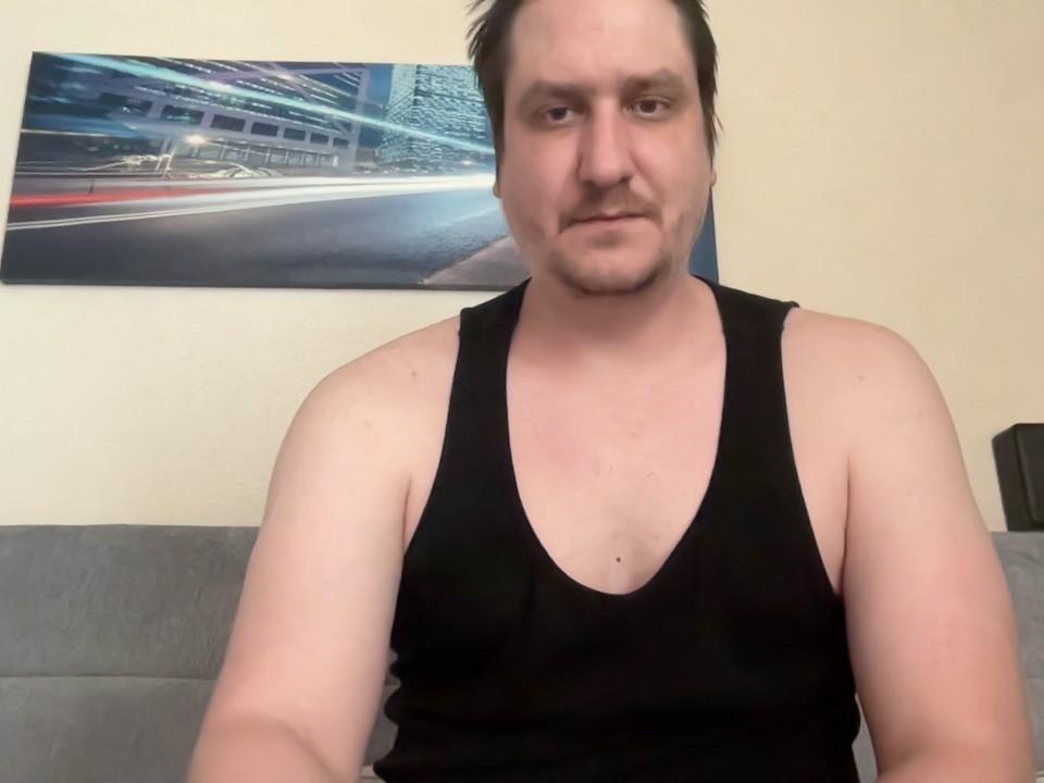 Alhem has a chubby face, short brown hair, and is wearing a black tank top, showing his tits and arms, sitting on a grey couch, looking slightly bored.