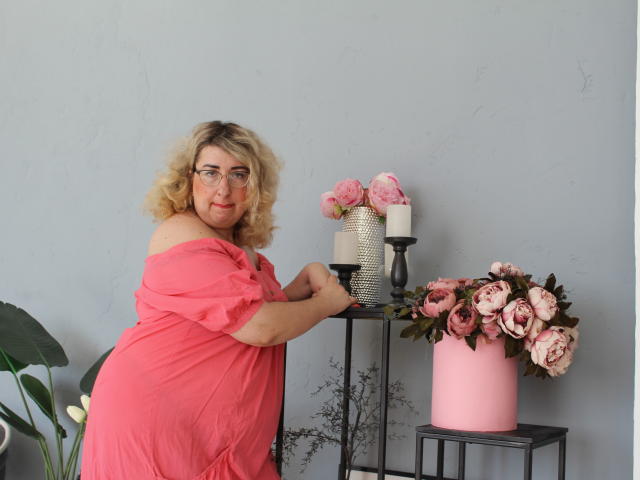 HelenEnigma, mit lockigem, blondem Haar und Brille, trägt ein pinkes Kleid mit einem Schulterauschnitt und steht neben einem schwarzen Metalltisch mit pinken Rosen, Kerzen und einer pinken Vase..