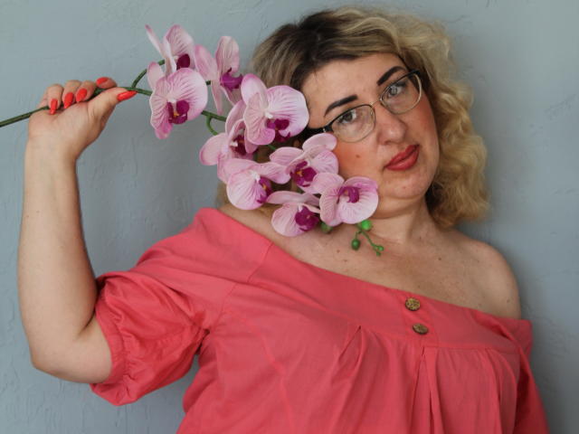 HelenEnigma with curly blonde hair, glasses, and red lipstick, holding pink orchids, wearing an off-shoulder pink top, showing her tits, against a light blue wall.