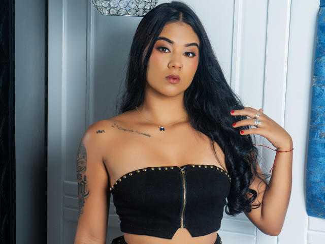 TamaraSummer with long black hair, tan skin, and full lips, wearing a black strapless top with gold studs, showing her small tits and tattoos, and red nails.