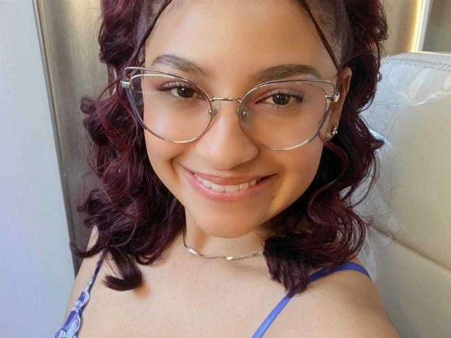Ameliebx tem cabelos ruivos escuros e cacheados, usa óculos de grau e tem um sorriso adorável. Ela está vestindo uma blusa azul com alças finas e um colar de prata. Seus seios são pequenos..