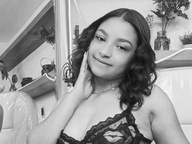 Ameliebx, avec ses cheveux noirs bouclés, un sourire en coin, touche sa joue, et porte de la lingerie en dentelle noire qui révèle ses seins. Noir et blanc, main sur le cou, sourire subtil..