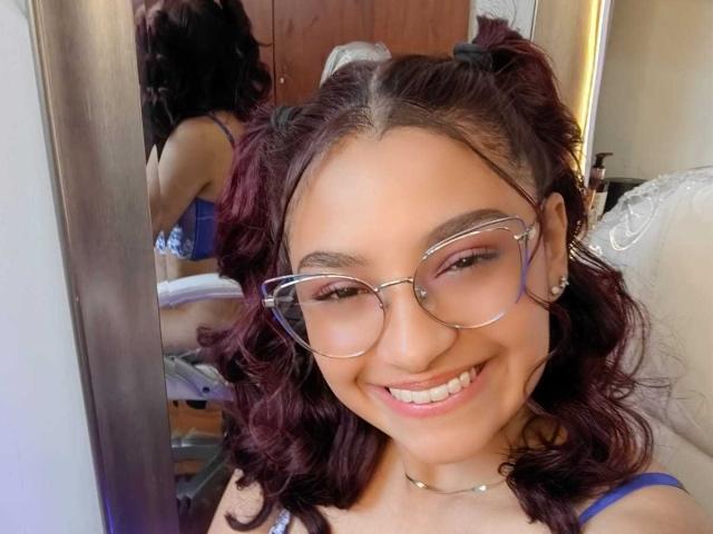 Ameliebx, com cabelo cacheado roxo escuro, sutiã azul e óculos, sorrindo em uma selfie no espelho, mostrando seu rosto fofo e dentes brilhantes, com o reflexo de sua bunda no espelho..
