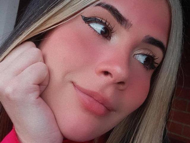 KrystinBrowm's face, showing pink blush, long blonde and black hair, thick eyebrows, long eyelashes, slight smile, hand resting on cheek, pink lips, looking to the side, soft pink skin.