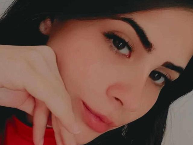 KrystinBrowm's face with dark eyebrows, black eyeliner, and pink lips, her hand resting on her cheek, looking slightly to the side with a subtle smile. Her skin is fair.