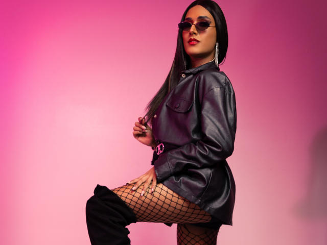 FallonRogers with long black hair, wearing sunglasses, black leather jacket, fishnet stockings, and black thigh-high boots, showing her ass and thigh in a pink-lit setting.