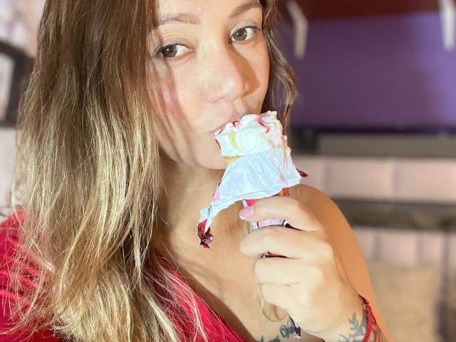 SophieLombardo with long brown hair, red shirt, eating a waffle cone with pink and white icing, looking at the camera, showing her pink-nail-painted hand, and a tattoo on her wrist.