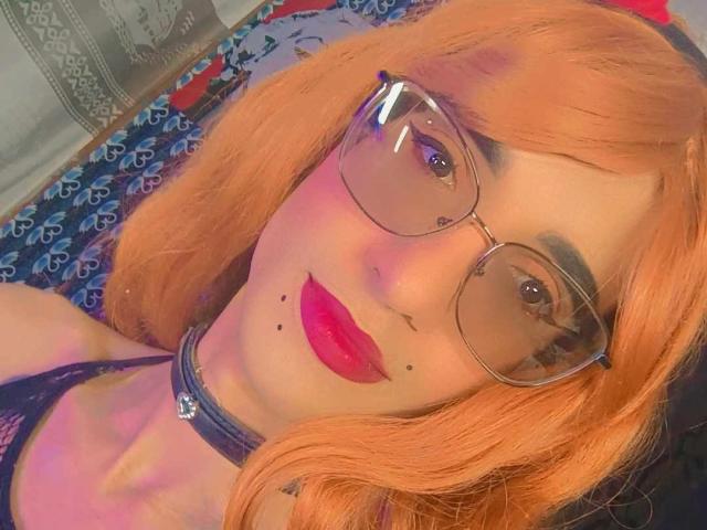 KrissiriWat with orange hair, glasses, red lipstick, and a black choker, showing a cute face with small black moles on her cheek. Boobs partially visible in lace top.