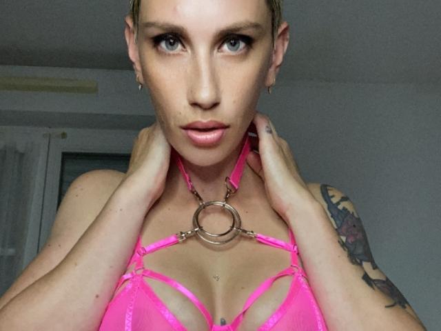 LizaRosa with short blonde hair, blue eyes, and pink lingerie with a large metal ring. She holds her collar, showing tattooed arm and small nipple piercings.