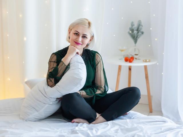 Olivia Horst, com cabelo loiro, usando uma blusa verde transparente, calças pretas, sentada em uma cama branca, segurando um travesseiro branco, sorrindo, com luzes suaves, uma mesa de madeira com copos de vinho, maçãs e uma planta..