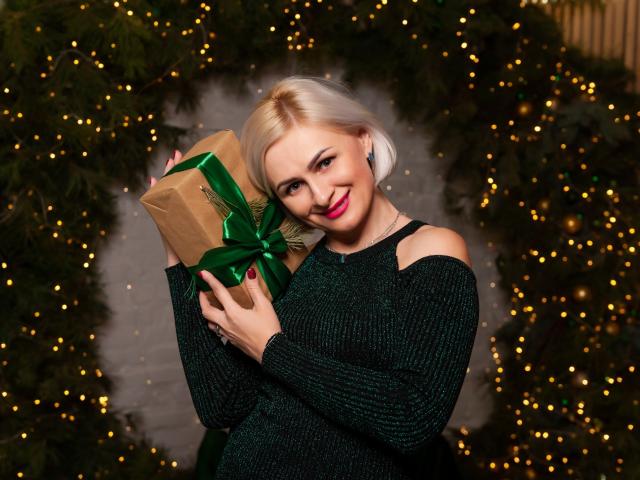 OliviaHorst with blonde hair, green sweater, holding brown gift with green ribbon, smiling, red lipstick, cute, Christmas tree lights in background, her tits slightly visible under sweater.