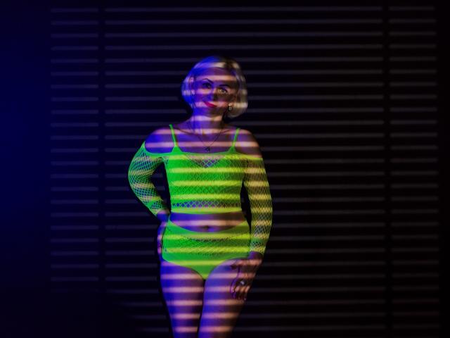 OliviaHorst wearing a neon green fishnet long-sleeve top and matching panties, standing with hands on hips, striped shadows across her body, blue and purple lighting.