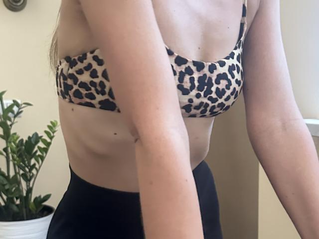 Stefanni's leopard-print bra, showing her small tits and flat stomach, with black pants and pale skin, leaning forward, arms in frame, potted plant on left.