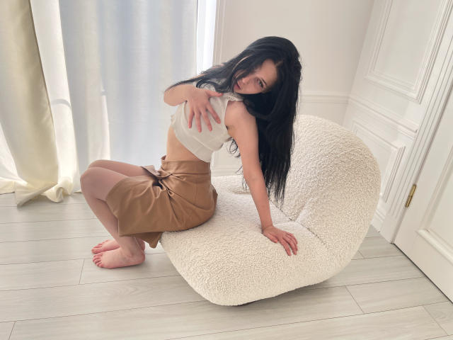 LiyaFrench with long black hair, tan skirt, and white top, sitting on a fluffy white chair, touching her small tits with a cute smile. Barefoot, light wood floor.