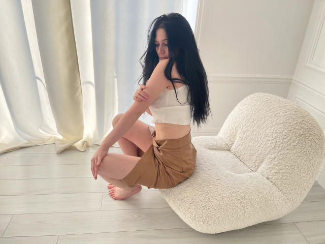 LiyaFrench with long black hair, white crop top, and tan skirt, sitting on a fluffy white chair, arms crossed, looking over shoulder, bare feet on light wood floor.