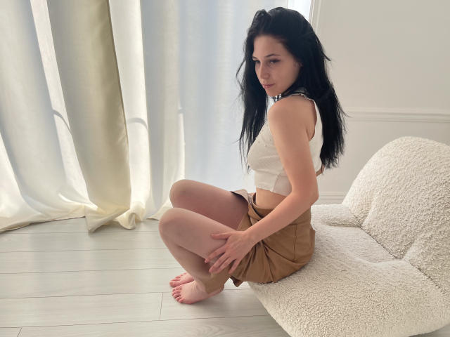LiyaFrench with long black hair, small white crop top, and brown skirt, squatting on a white textured chair, barefoot, hands on knees, looking over shoulder.