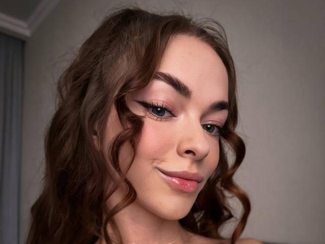 UnderTheSkin tem cabelo castanho ondulado, sobrancelhas grossas, delineador de olhos e um sorriso discreto. Seus lábios rosados estão levemente brilhantes. Ela está olhando para o lado com uma expressão adorável..