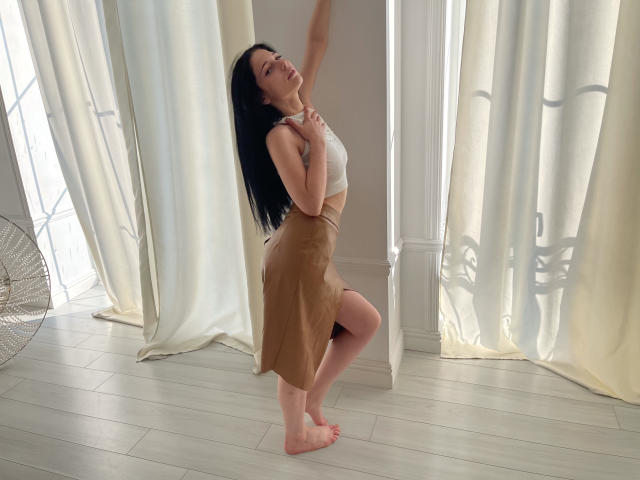 LiyaFrench, barefoot, in a white crop top, tan skirt, standing on one leg, arms raised, black hair, light skin, wooden floor, white curtains, window, fan on left.