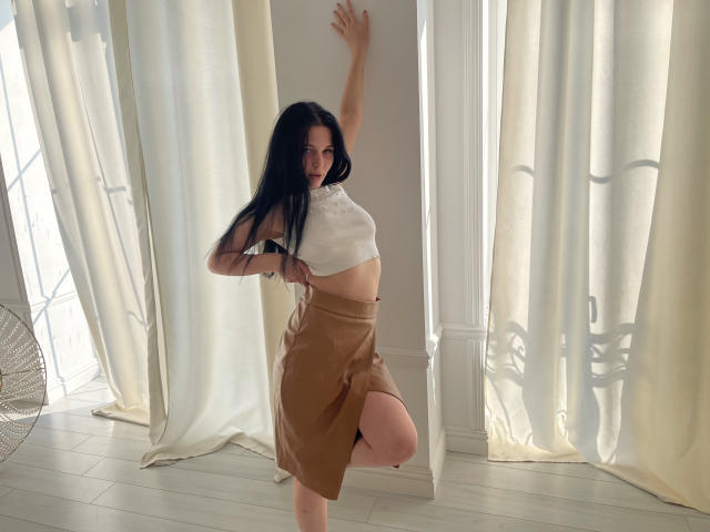 LiyaFrench with long black hair, white sleeveless top, and brown skirt, standing on one leg, arm raised, exposing her tits and showing her ass. Sunlight filters through curtains.