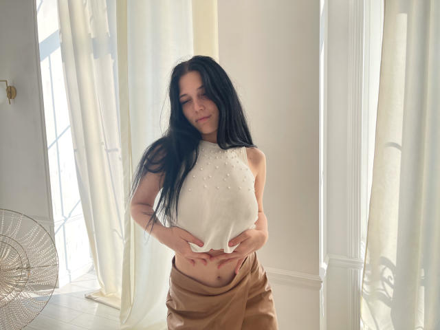 LiyaFrench with long black hair, white sleeveless top with studs, pulling down a beige satin skirt, showing her bare belly and hands. Soft smile, natural light.