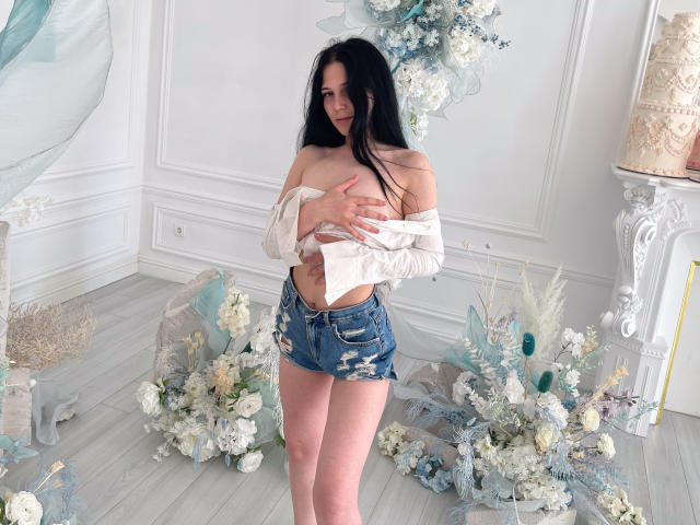 LiyaFrench with long black hair, off-shoulder white top, and torn blue denim shorts, covering her small tits with her hand, standing in a room with floral arrangements.