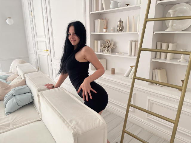 LiyaFrench with black hair, wearing a tight black dress, leaning on a white couch, showing off her ass, smiling, standing in a bright, white room with a gold ladder.