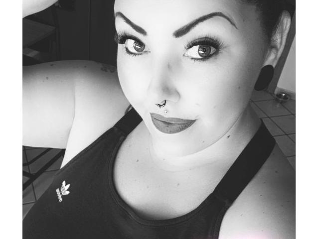 TinyDiamond88-hot a des sourcils foncés, un piercing au nez et du rouge à lèvres foncé. Elle porte un débardeur noir avec un petit logo, et a de grands yeux ronds qui regardent la caméra..