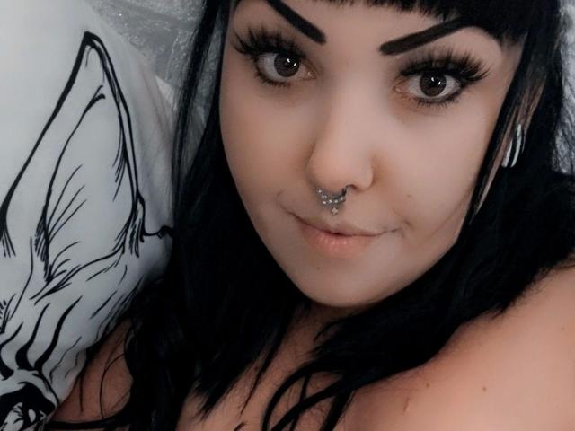 TinyDiamond88 - aantrekkelijk met zwart haar, dikke wenkbrauwen, lange wimpers, een neusring, een septum piercing en een lichte glimlach, kijkt naar de camera, ligt op haar rug met een kussen..