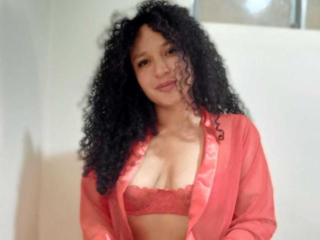CarlaMuille with curly black hair, red lace bra, and matching red satin robe, standing, showing her small tits, smiling softly, light brown skin, light background.