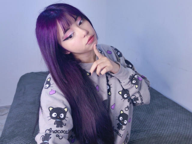 Arieyaara, con lunghi capelli viola, eyeliner rosa e un maglione grigio a forma di gatto, con un dito sulle labbra, sguardo seducente, con un letto grigio dietro di lei. Il seno non è visibile..