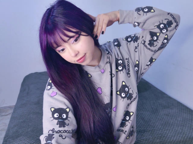 Arieyaara, avec de longs cheveux violets, portant un sweat à capuche gris en forme de chat, assise sur un lit vert, une main dans les cheveux, souriant doucement. Les seins ne sont pas visibles, l'accent est mis sur le visage et le sweat à capuche..