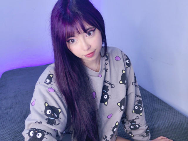 Arieyaara mit langen, violetten Haaren, die ein graues Pyjamaoberteil mit Katzenmuster trägt, sitzt auf einem Bett und blickt mit einem leichten Lächeln in die Kamera. Im Hintergrund ist violettes Licht zu sehen..