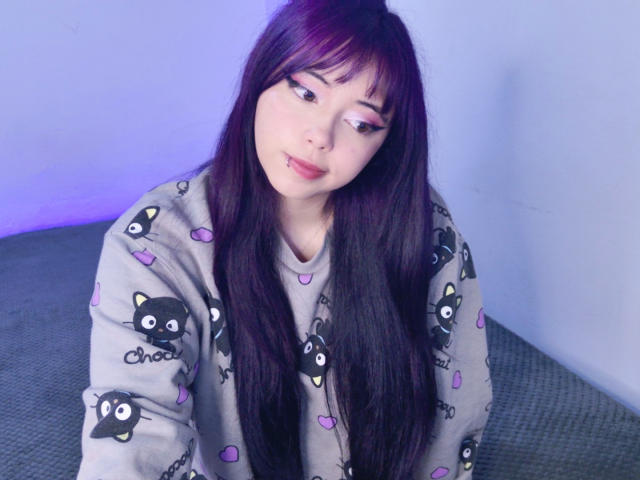 Arieyaara with long purple-tinted black hair, wearing a gray cat print shirt, has a slight pout and looks down with pink eyeliner and a nose ring.