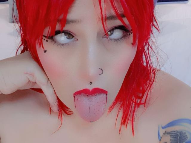 IsabellaRoshe with bright red hair, green eyes, nose piercings, and a lip piercing, sticking out her tongue. She has pink blush, red lipstick, and a tattoo on her shoulder.