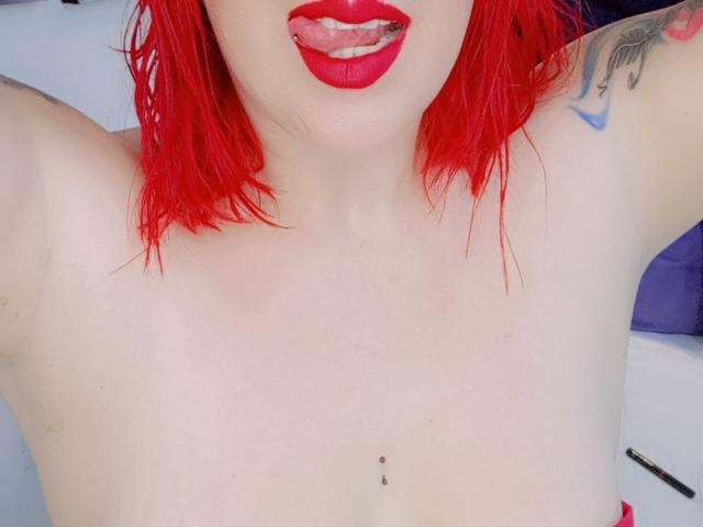 IsabellaRoshe with bright red hair, red lipstick, and tongue out, showing a nipple piercing and a tattoo on her right arm. Boobs, tits, and a small black stud.