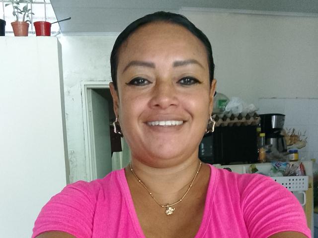 Crysmia has brown skin, black hair in a ponytail, small tits, wearing a pink shirt, gold necklace, and earrings, smiling with white teeth in a selfie.
