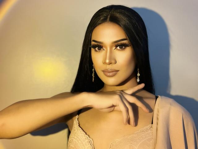 NeedGoodSweetie with long black hair, wearing lace top, has tan skin, full lips, and is pointing with her right hand, wearing dangling earrings, and has thick, dark eyebrows.