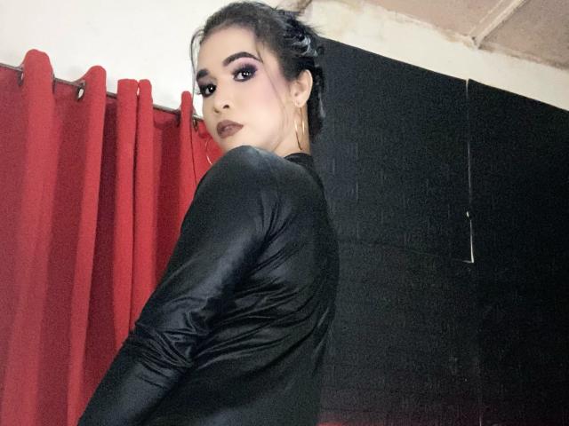 LexiSmithX has dark makeup, a black shiny long-sleeve top, and a side glance, showing off her ass. Red curtains and black wall are in the background.