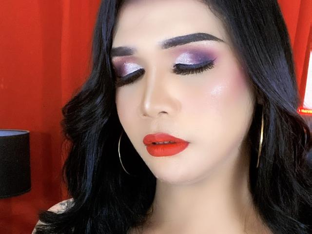 LexiSmithX hat glänzenden, violetten Lidschatten, roten Lippenstift und schwarzes, welliges Haar. Sie trägt goldene Ohrringe im Stecker-Stil und hat die Augen geschlossen, wobei ihre langen, dunklen Wimpern sichtbar sind..