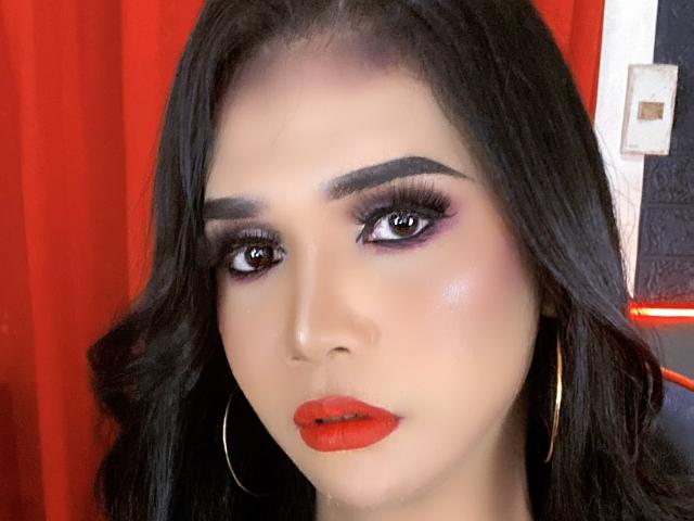 LexiSmithX hat dunkles, welliges Haar, dicke Augenbrauen, lila Lidschatten, lange Wimpern und leuchtend roten Lippenstift. Sie trägt goldene Ohrringe im Stecker-Stil und blickt direkt in die Kamera..
