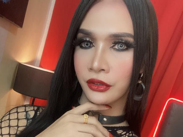 LexiSmithX has long black hair, blue eyes with thick eyelashes, red lipstick, and is wearing a black fishnet top with a black choker. Her hand is near her chin.