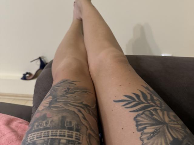 AndraD's tattooed legs, crossed at the ankles, showing detailed black ink tattoos of a building and palm leaves on her thighs, with her bare feet in the background.
