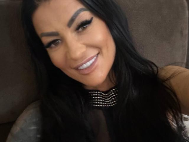AndraD has long black hair, thick eyebrows, and a big smile showing white teeth. She's wearing a black top with a spiked collar, looking cute and sexy.