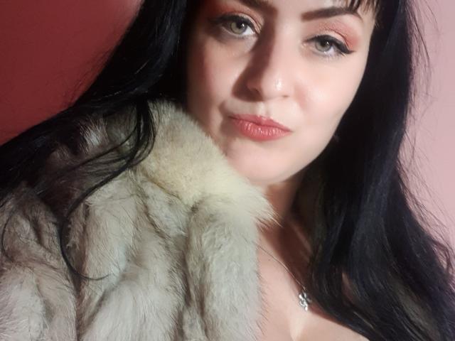 MistressMayaDiamondXx with black hair, pale skin, and pink lipstick, wearing a furry grey coat, showing her tits slightly, and a silver necklace with a small pendant.