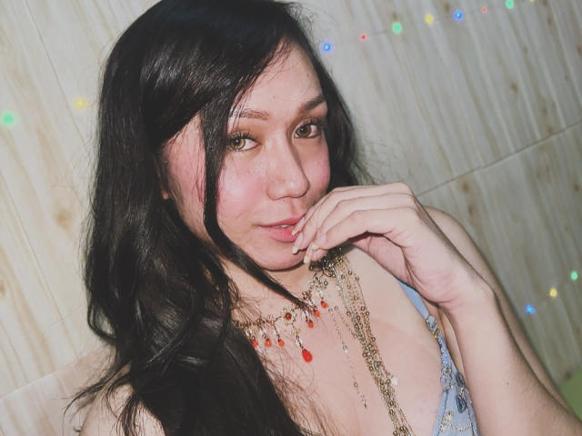 TsJade with long black hair, brown eyes, and a slight smile, biting her finger, wearing a blue lace top and red bead necklace, showing her tits.